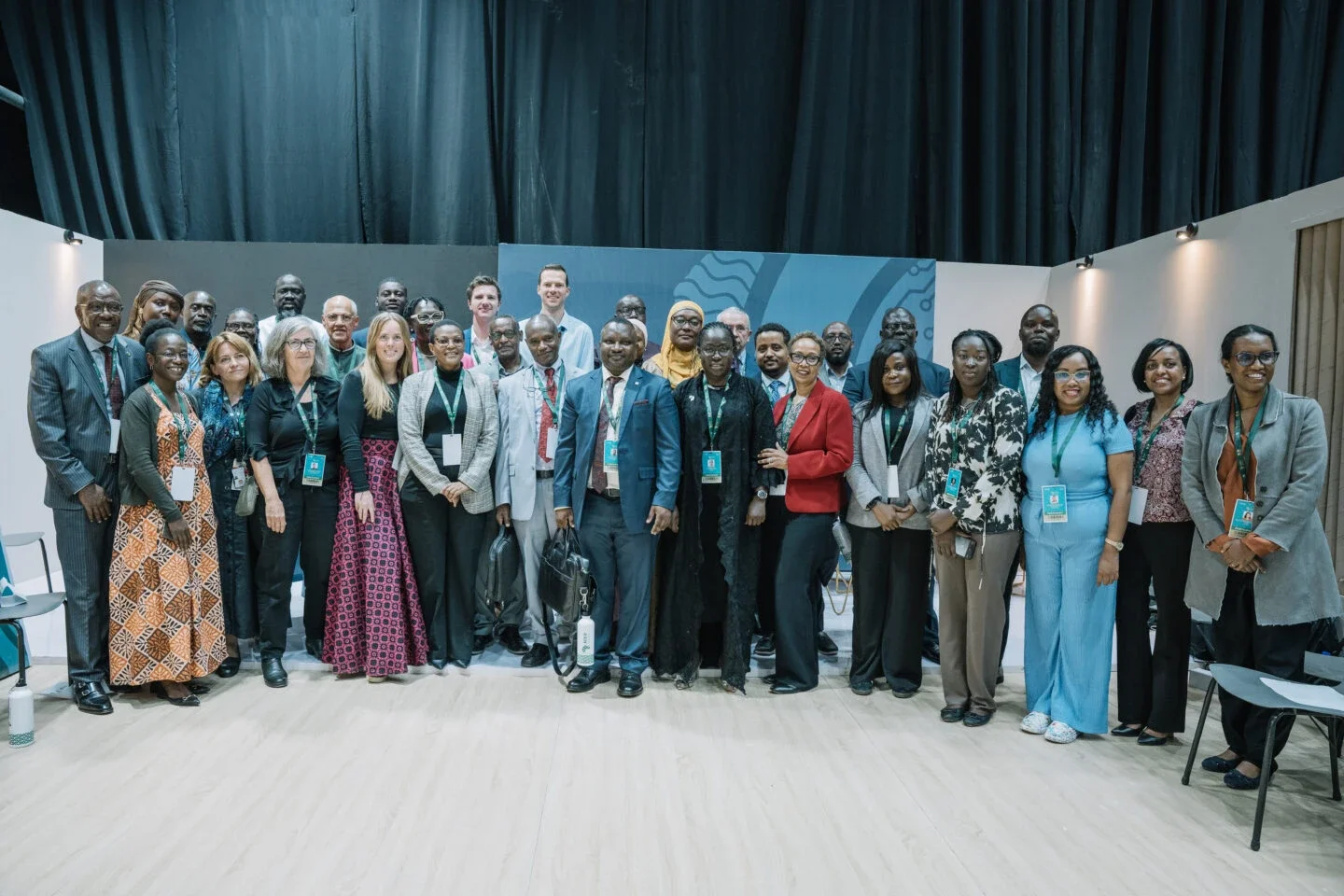 large group of conference participants posing together indoors, with many wearing badges and lanyards, smiling at the camera.