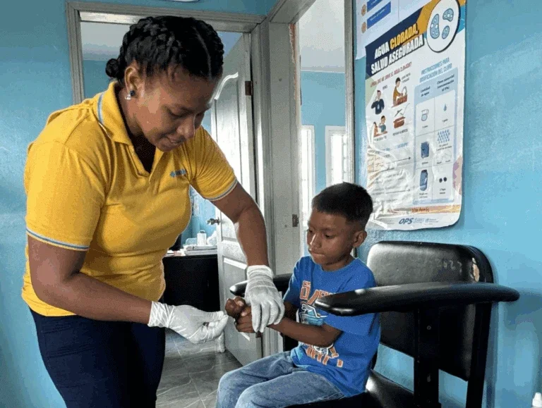 a healthcare worker wearing gloves checks a young boy’s hand in a clinic, with a health poster about safe water on the wall behind them.