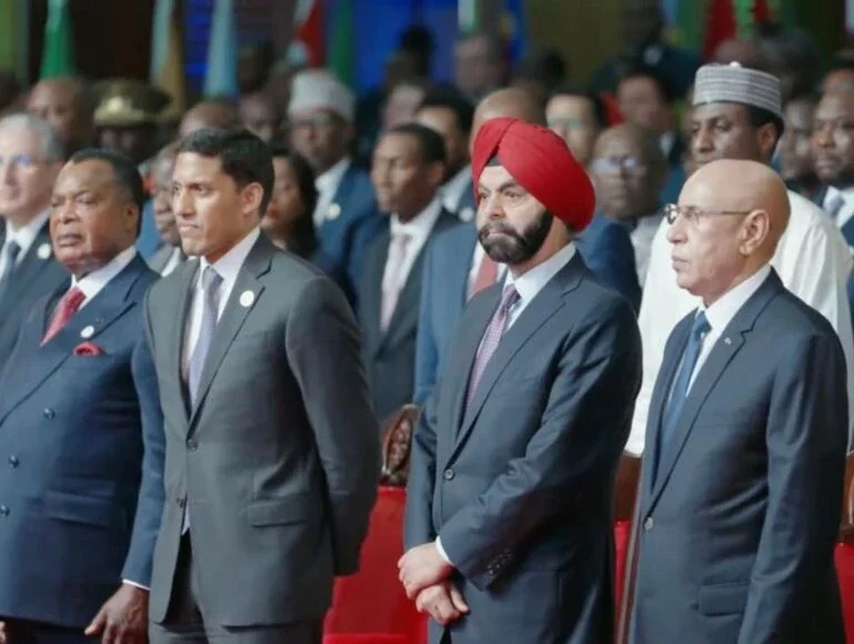 roup of men in suits standing together during a formal international event, with flags and a large audience visible in the background.